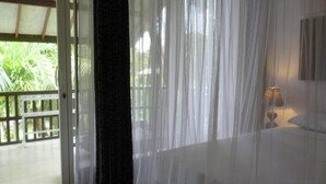 Una habitación con cama, ventana con cortinas translúcidas y vista a la vegetación.