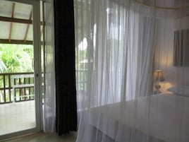 Una habitación con cama, ventana con cortinas translúcidas y vista a la vegetación.