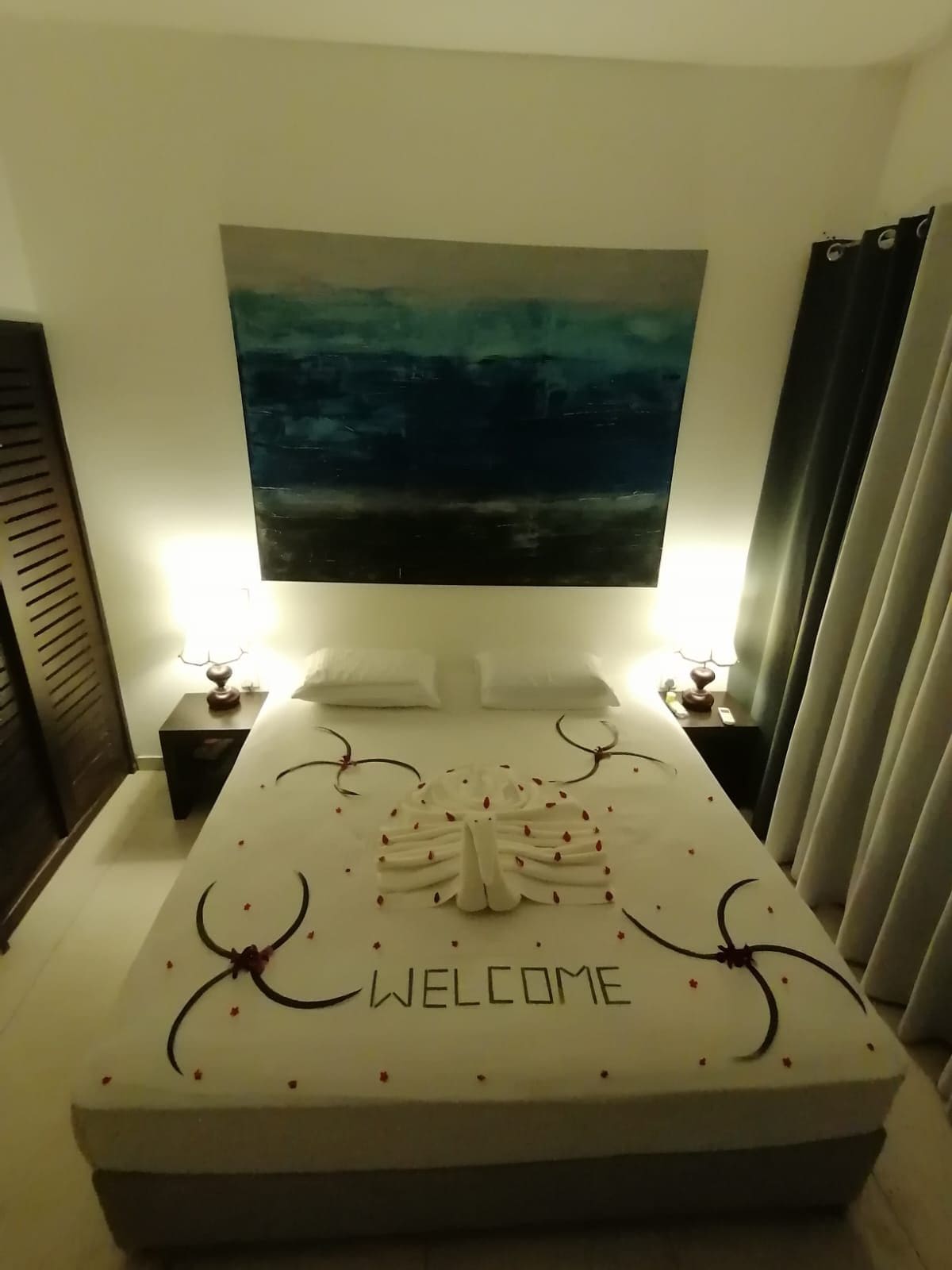 Un lit avec une pancarte « BIENVENUE », un grand tableau au-dessus et une table de chevet avec une lampe.