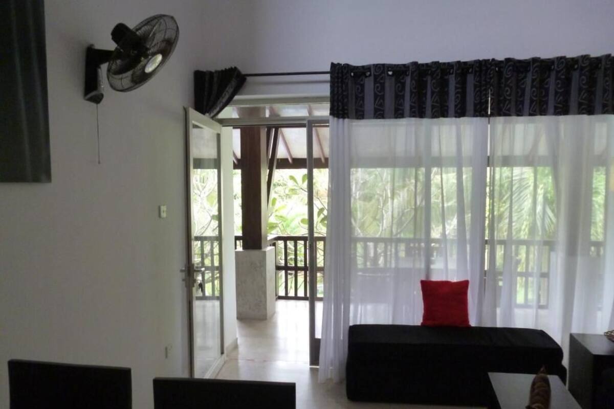 A room with a black sofa, a small table, a wall-mounted fan, and a large window with sheer curtains.