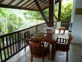 Un patio al aire libre con una mesa y sillas de madera, rodeado por una baranda negra, con vistas a un exuberante jardín.