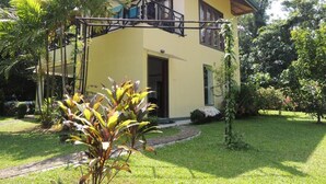 Una casa de dos pisos con un balcón, rodeada de un jardín cuidado y árboles.