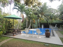 Zona junto a la piscina con una mesa y sillas, rodeada de palmeras y una pequeña estatua.