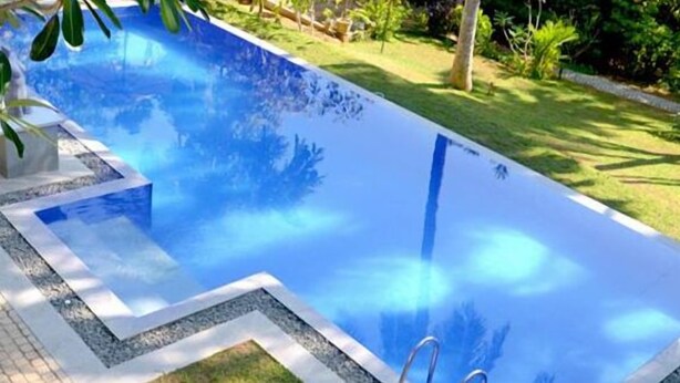 Una piscina rectangular con superficie de agua azul cristalina, rodeada por un camino pavimentado y abundante vegetación.