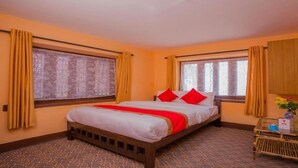A hotel room with a bed, two windows with curtains, a television on the wall, and a small table with a vase of flowers. - MeroStay 154 White & Peace (Lalitpur)