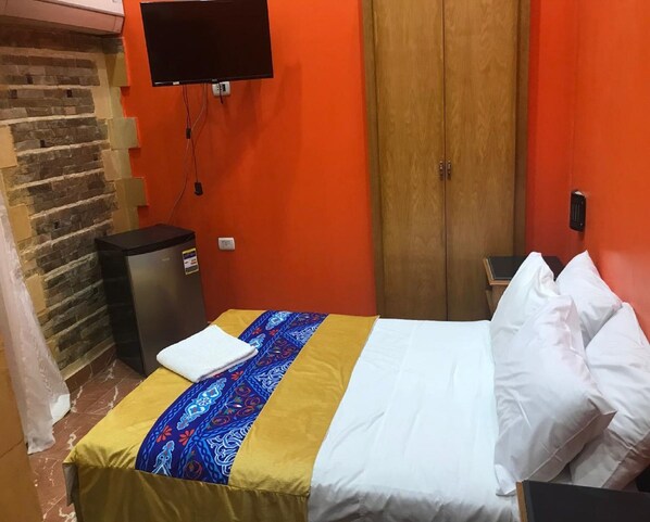 A hotel room with a bed, a television mounted on the wall, a wooden wardrobe, and a small trash bin.
