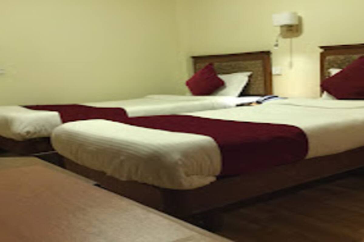 A hotel room with two single beds, each with a red pillow and white bedding. There is a wooden nightstand with a lamp on it.
