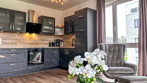 A modern kitchen with dark cabinets, a built-in oven, and a flat-screen TV. There is a grey armchair and a vase of white orchids by the window. - Weiße Villen Wohnung 04 (Ostseebad Boltenhagen)