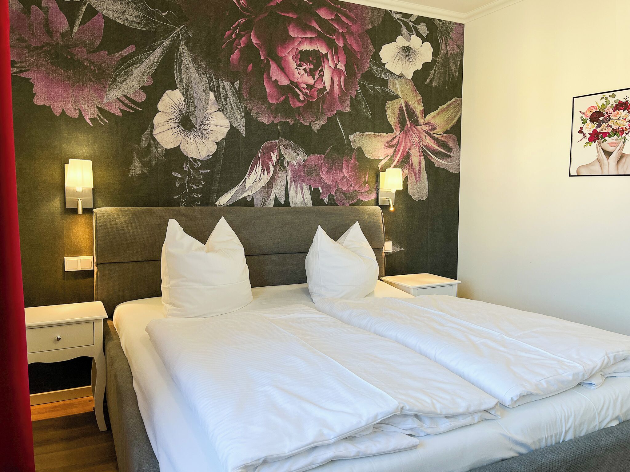 A hotel room with a large bed, two bedside tables, a nightstand, a red curtain, and a floral wall mural.
