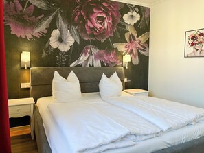 Una habitación de hotel con una cama grande, dos mesitas de noche, una lámpara de noche, una cortina roja y un mural floral en la pared.