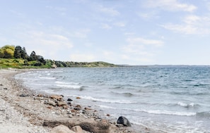 Une plage de galets et de rochers, une mer calme et, au loin, un paysage vallonné.