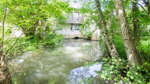 Un cadre paisible au bord de la rivière, avec un pont de pierre rustique, une végétation luxuriante et un bâtiment doté d’un balcon donnant sur l’eau.