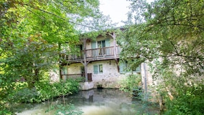 Un bâtiment rustique en pierre, doté d’un balcon, entouré d’une végétation luxuriante et d’un petit étang.