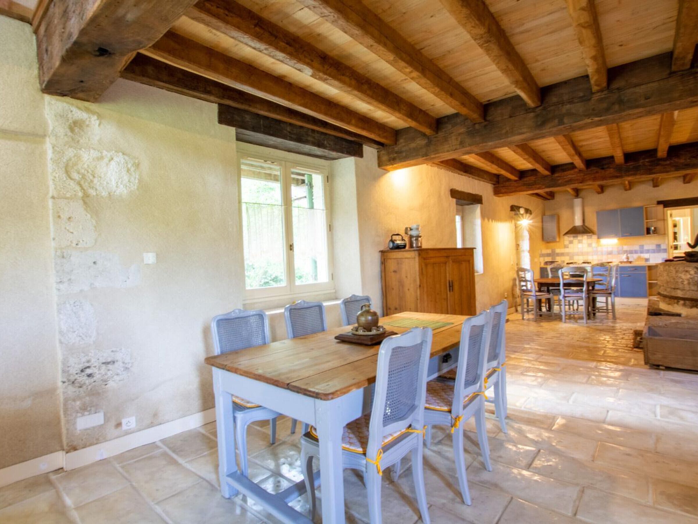 A rustic dining area with a wooden table and chairs, a kitchen in the background, and a stone floor.