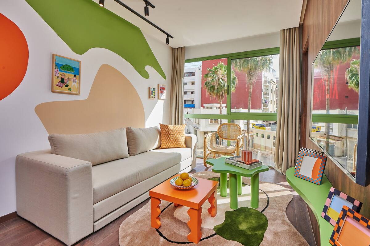 Una sala de estar moderna con un sofá, una mesita baja con un cuenco de frutas, una mesita auxiliar verde y un ventanal con vistas a edificios y palmeras.