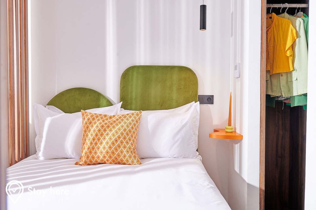 Una cama bien hecha con almohadas blancas y estampadas, un cabecero verde y una mesita de noche naranja con una lámpara.