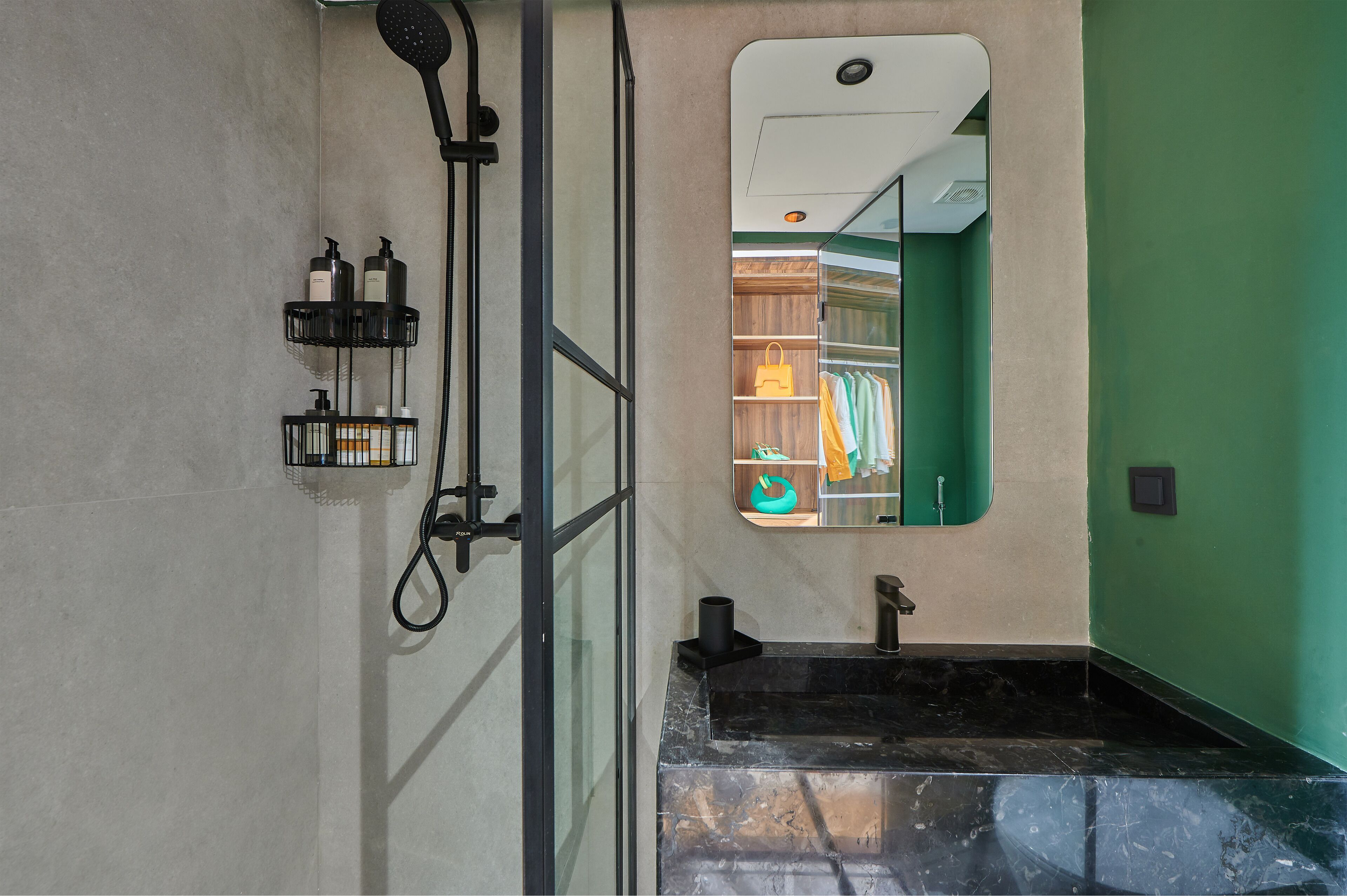 A bathroom with a black marble sink, a shower with a handheld showerhead, and a mirror reflecting a green wall and a closet with clothes.