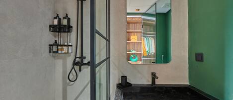 A bathroom with a black marble sink, a shower with a handheld showerhead, and a mirror reflecting a green wall and a closet with clothes.