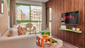 Una sala de estar moderna con un sofá, una mesita con un cuenco de fruta, una mesita auxiliar verde, un sillón de mimbre y un televisor grande montado en la pared.
