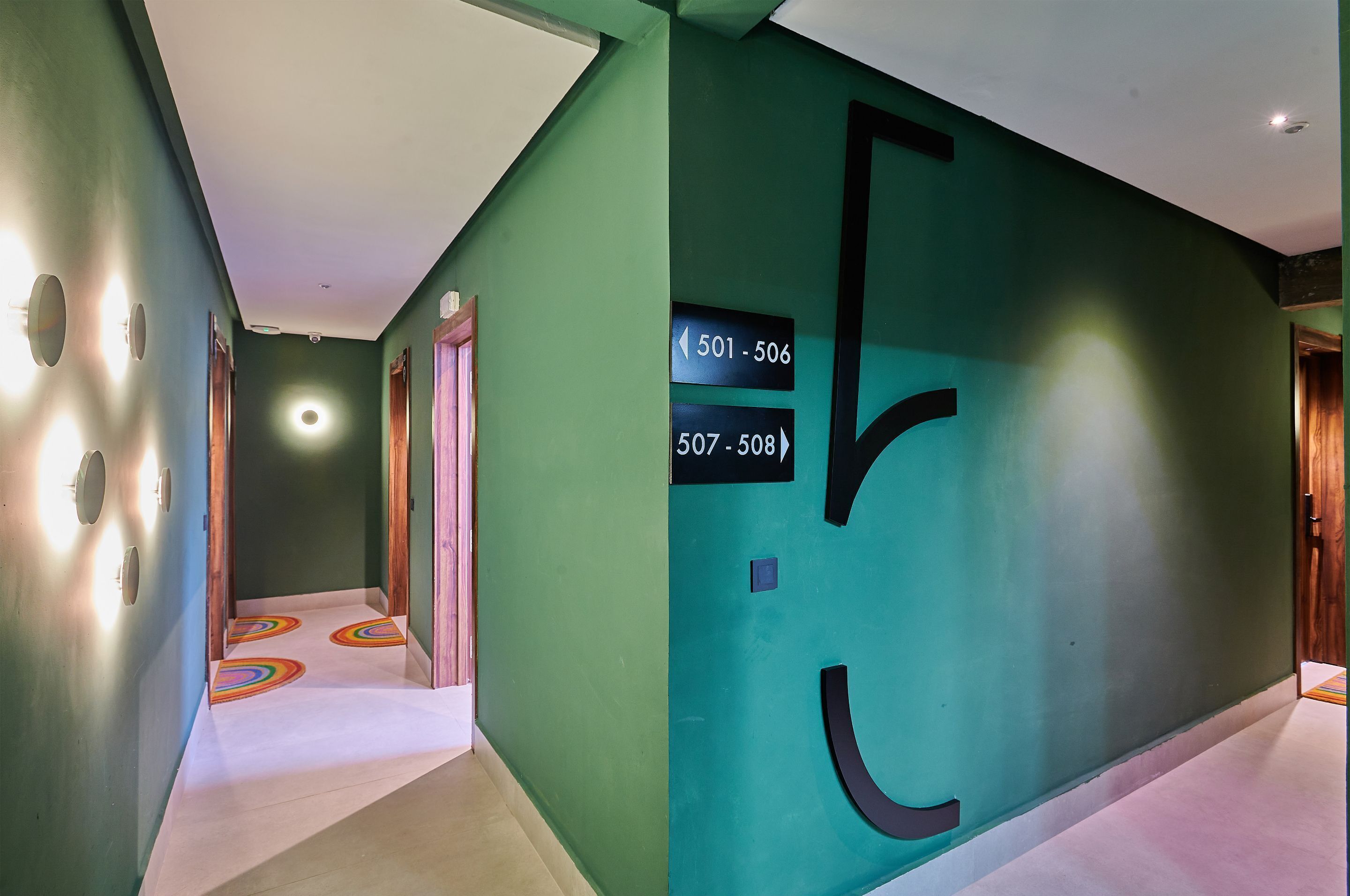 Un couloir aux murs verts, avec des portes numérotées et un luminaire circulaire.