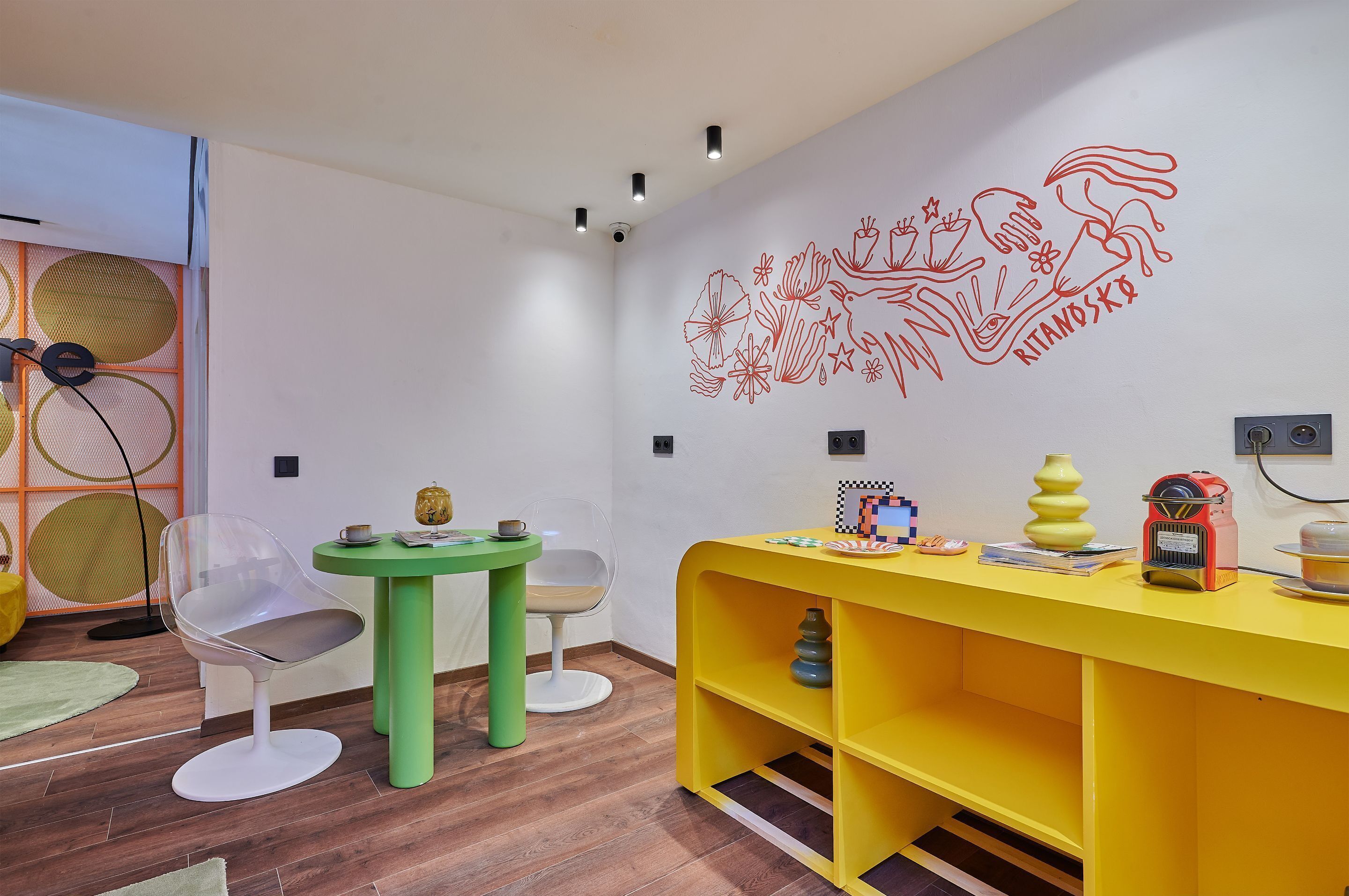 A room with a yellow desk, a green table, and a clear chair. There is a wall with red drawings and a small shelf with a vase and a radio.