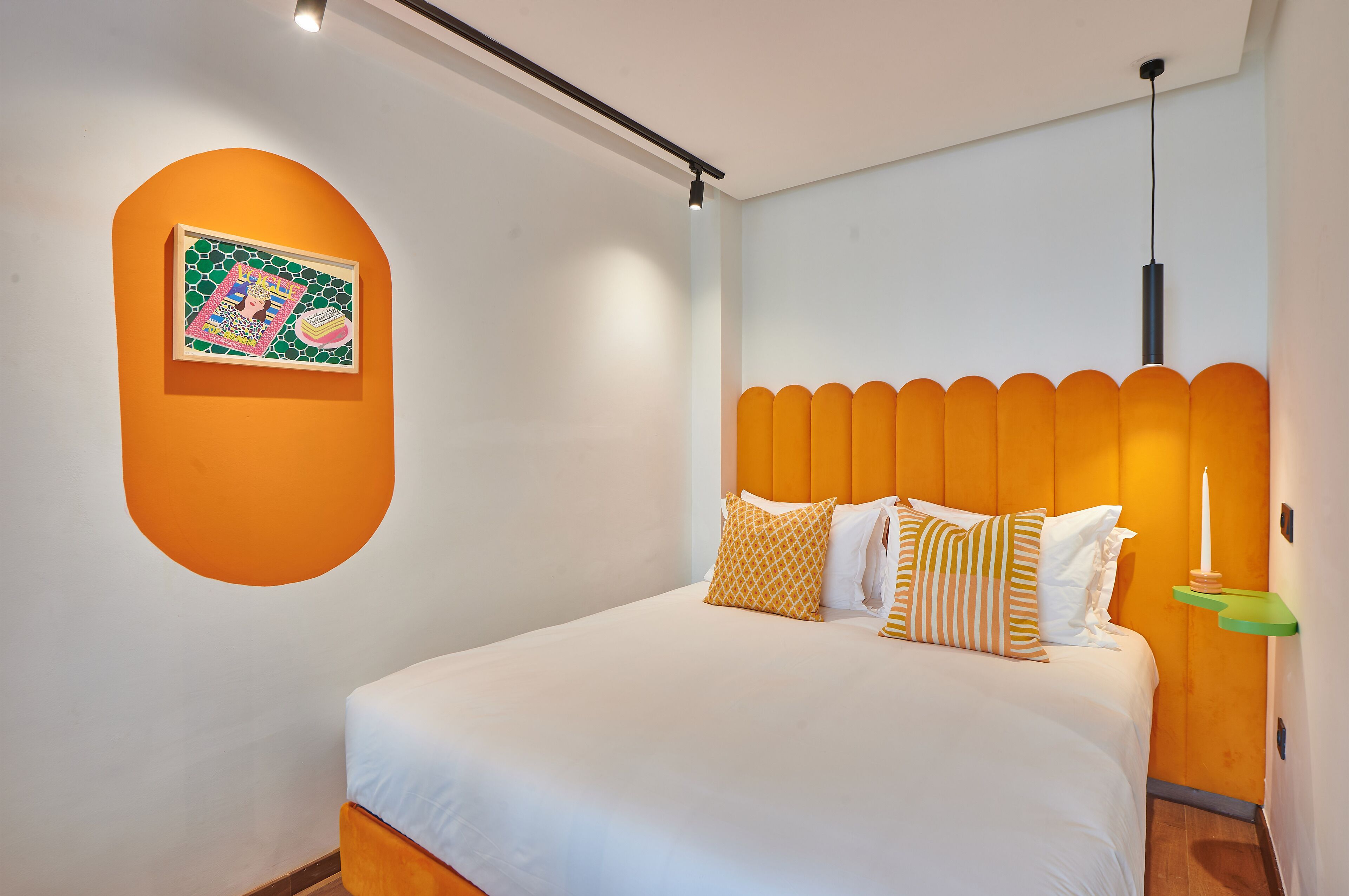 Una cama bien hecha con ropa blanca y varias almohadas, una obra de arte colgada en la pared y una pared de acento naranja única.