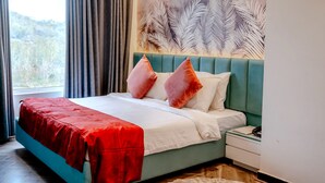 A modern bedroom with a teal bed, red and white bedding, a white nightstand, and a large window with curtains. - Redwood Resort (Panchkula)