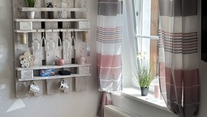 A corner of a room with a wooden shelf holding various items, a window with striped curtains, a radiator, and a wicker chair. - Büschem feel-good paradise (Lindlar)