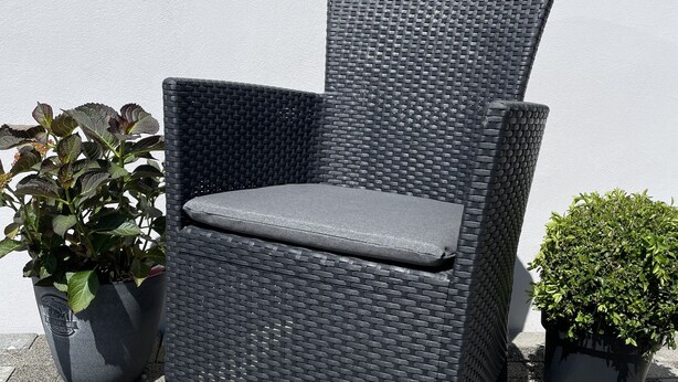 A wicker chair with a cushion, placed on a paved surface next to two potted plants. - Büschem feel-good paradise (Lindlar)
