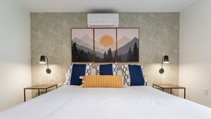 Ein ordentlich bezogenes Bett mit weißer Bettwäsche und einem zentralen gelben Kissen, umrahmt von blauen und weißen Kissen mit Muster. Zwei Nachttische mit Lampen, und eine wandmontierte Klimaanlage über einem gerahmten Landschaftsbild.