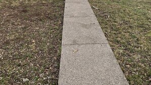 Ein Wohnhaus mit Vorgarten, einem Betonweg, der zur Haustür führt, und einer kleinen Veranda.