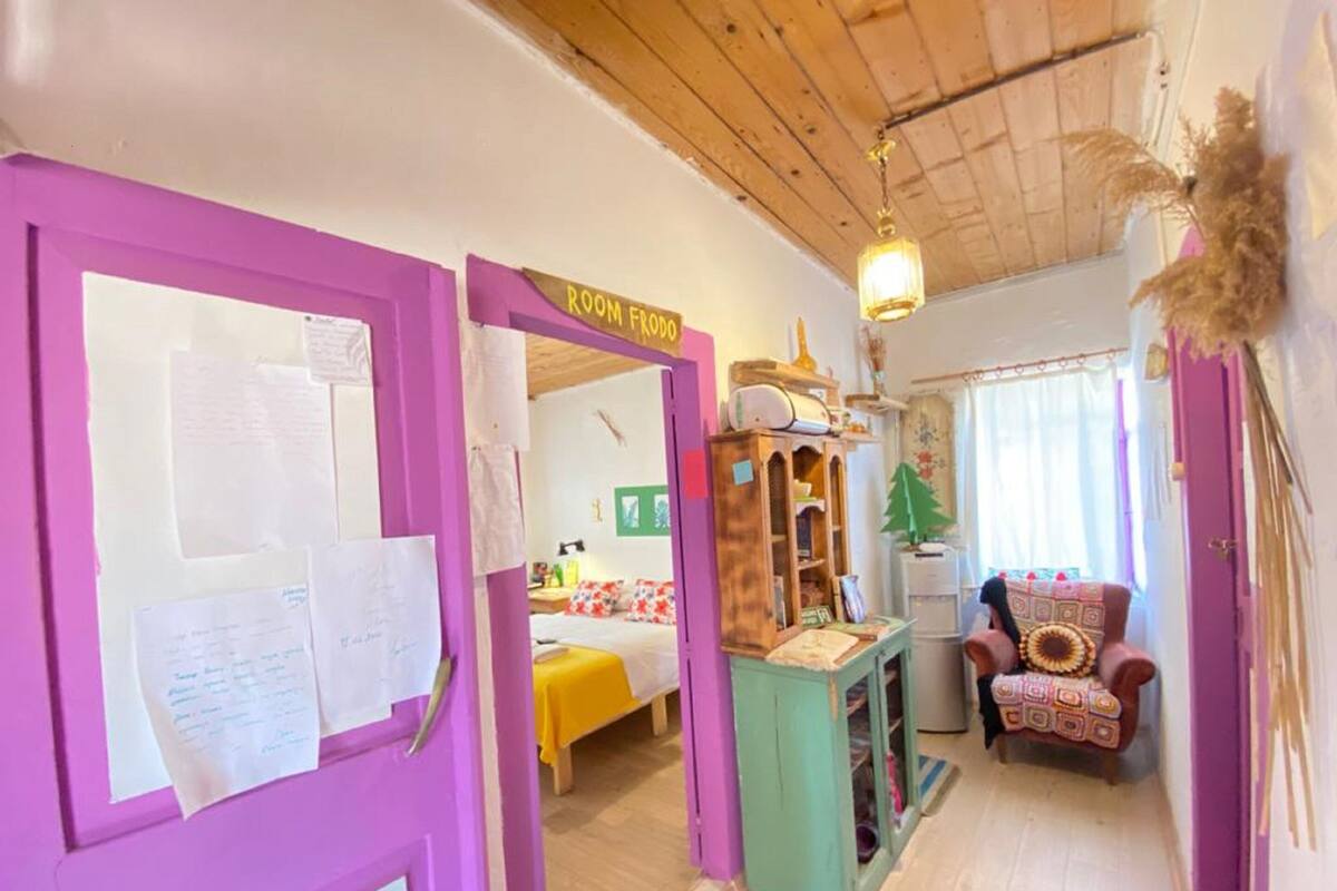 A room with a purple door, a bed with a yellow bedspread, a small wooden cabinet, and a chair with a patterned cushion.