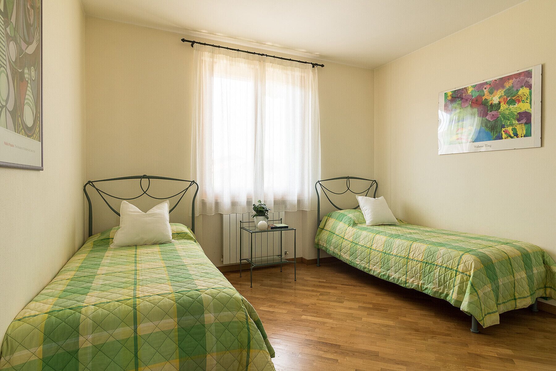 Un dormitorio con dos camas individuales, una ventana con cortinas traslúcidas, un radiador y un cuadro en la pared.