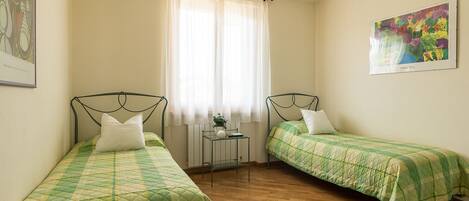 Un dormitorio con dos camas individuales, una ventana con cortinas traslúcidas, un radiador y un cuadro en la pared.