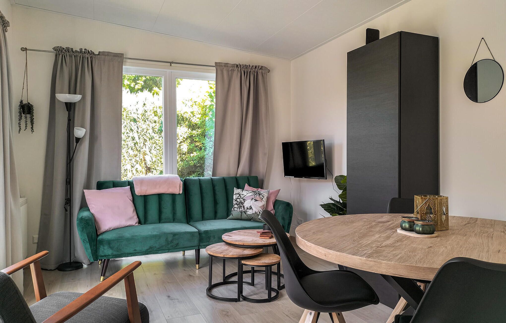 A modern living room with a green sofa, a round wooden dining table, and a flat-screen TV mounted on the wall.