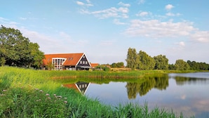 A traditional Dutch house with a red roof, situated beside a calm lake with lush greenery and trees in the background. - Stunning home in Heinkenszand with WiFi (Heinkenszand)