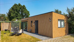A modern wooden cabin with a sliding glass door, a small patio area with a grill and lounge chairs, and a paved walkway. - Stunning home in Heinkenszand with WiFi (Heinkenszand)