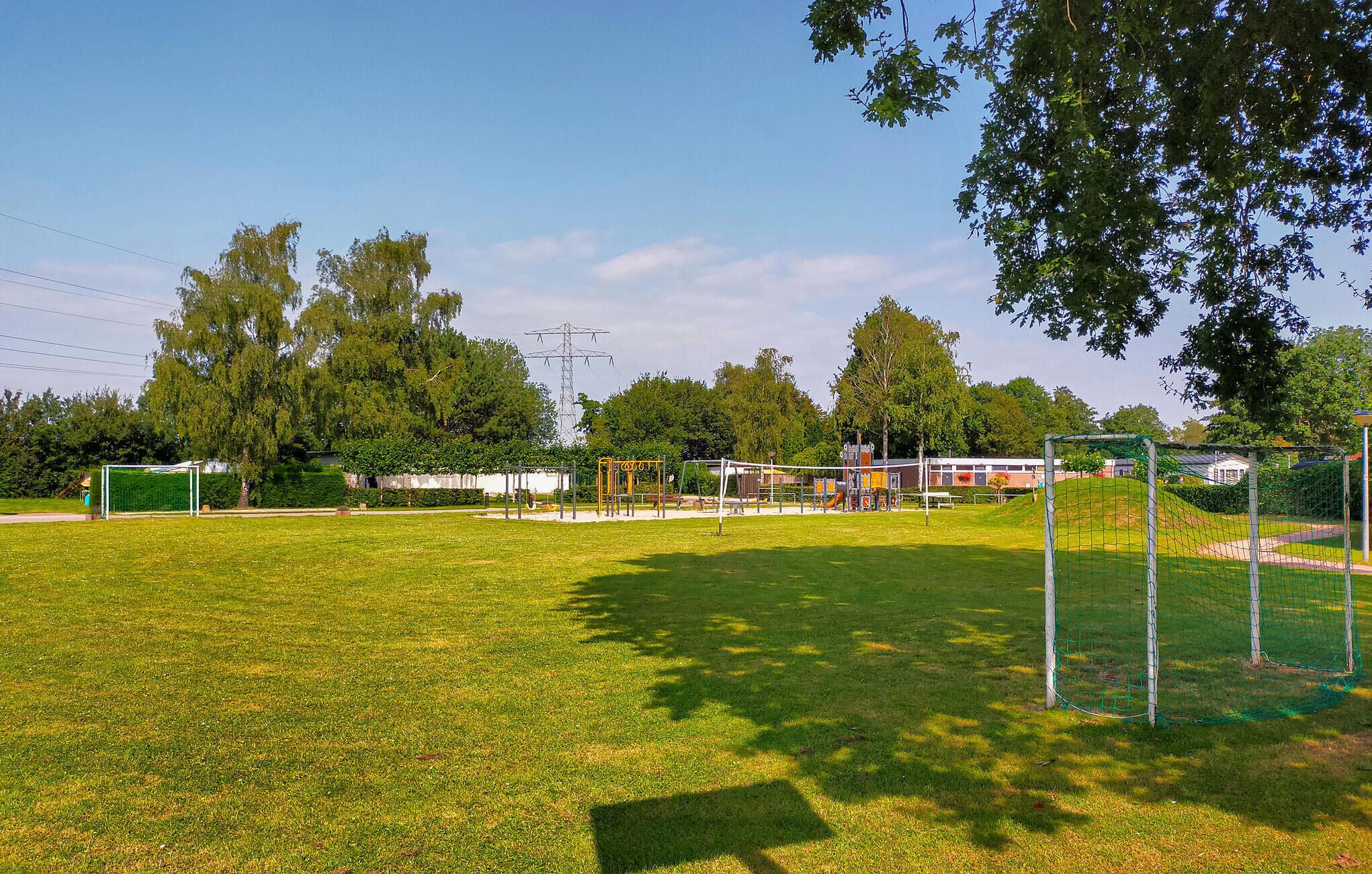A grassy field with playground equipment, a soccer goal, and trees.