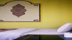 A single bed with a white mattress and pillow, a small bedside table with a tissue box, and a wall with a decorative pattern. - Merostay 158 Hotel RM (Kathmandu)