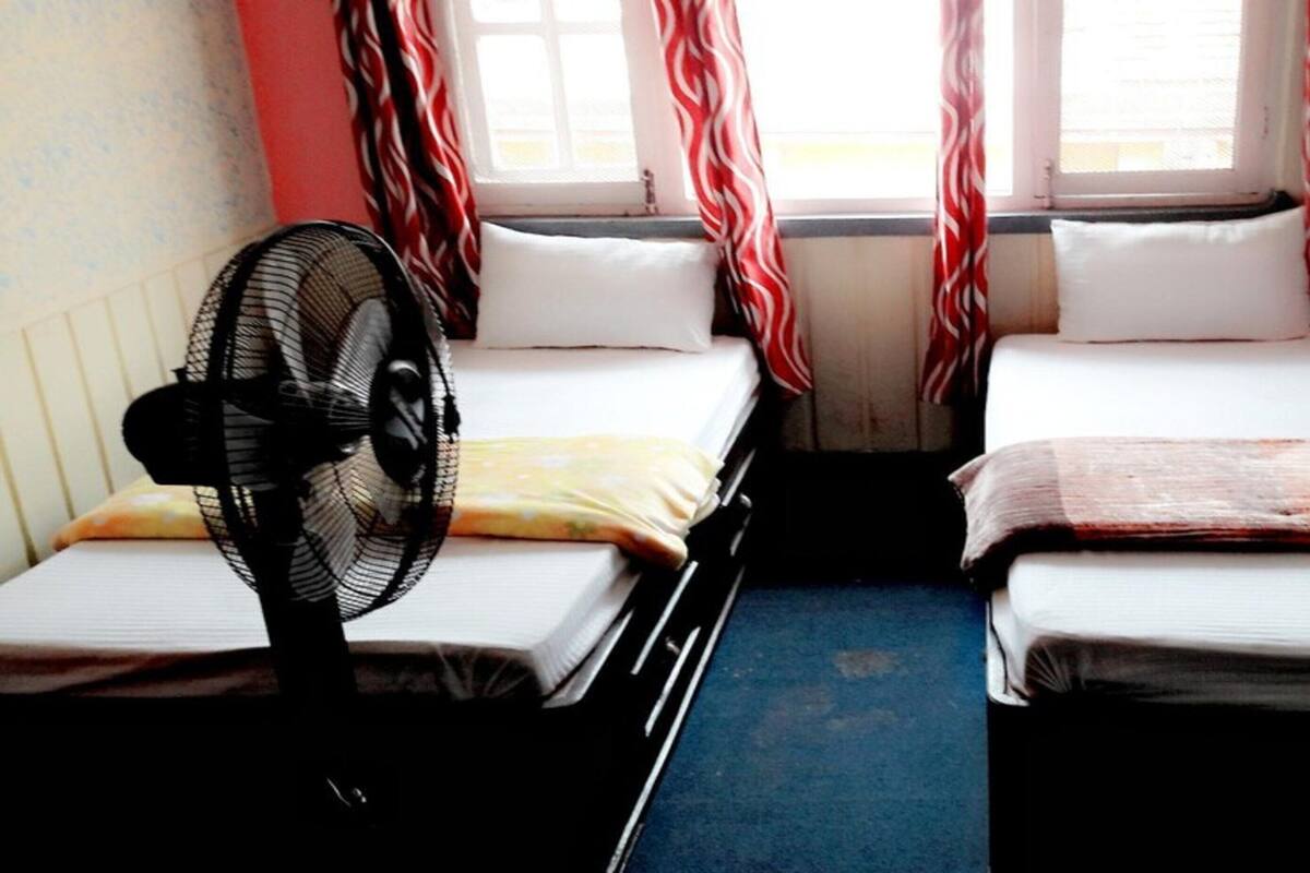 A small room with two beds, a standing fan, and a window with curtains.