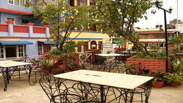 An outdoor patio area with tables and chairs, surrounded by potted plants and a building with a sign for a restaurant. - Merostay 158 Hotel RM (Kathmandu)