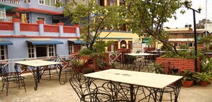 An outdoor patio area with tables and chairs, surrounded by potted plants and a building with a sign for a restaurant. - Merostay 158 Hotel RM (Kathmandu)