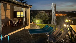 A terrace with lounge chairs, a table, and a view of a town at dusk. - Villa Crilu Sardinia, cozy, with sea view, swimming pool and private parking space (Tanaunella)