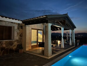 Ein überdachter Außenbereich mit Pool, Steinmauern und Blick auf die Berge in der Abenddämmerung.