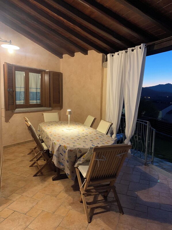 Un patio techado con una mesa redonda y sillas, una ventana con contraventanas y vistas al paisaje.
