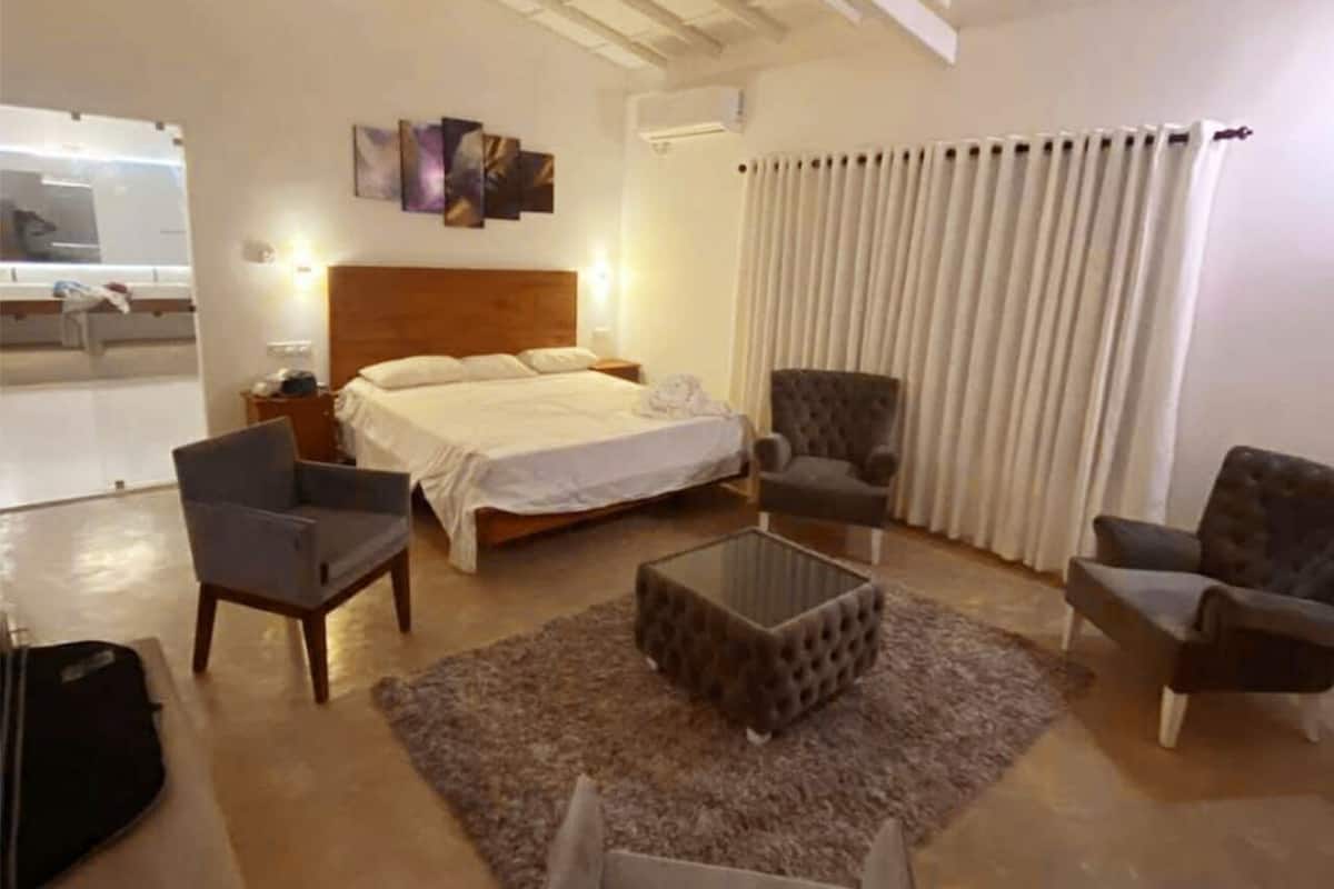 Habitación de hotel con cama, dos sillones, una mesita de centro y un ventanal con cortinas.