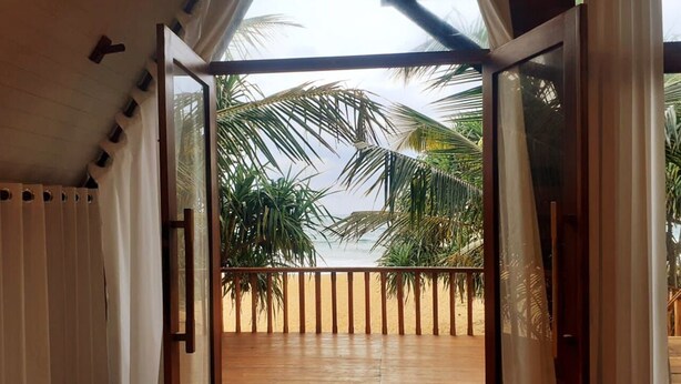 A room with a wooden floor, a sliding glass door leading to a balcony with a view of palm trees and a beach.