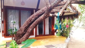 A house with a large tree branch overhanging the entrance, a thatched roof, and a paved patio area with potted plants.