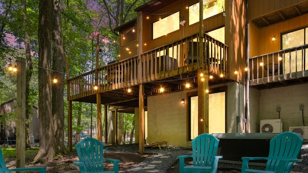 A two-story wooden cabin with outdoor seating and string lights. - Hot Tub, Fire Pit, Game Room; 4BR Cozy Poconos! (East Stroudsburg)