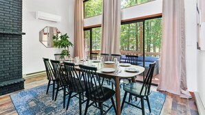 A dining area with a round table, black chairs, and a blue rug. There is a brick wall on the left and a large window with curtains. - Hot Tub, Fire Pit, Game Room; 4BR Cozy Poconos! (East Stroudsburg)